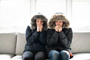 cold-couple-sitting-on-couch-in-heavy-parkas-with-hoods